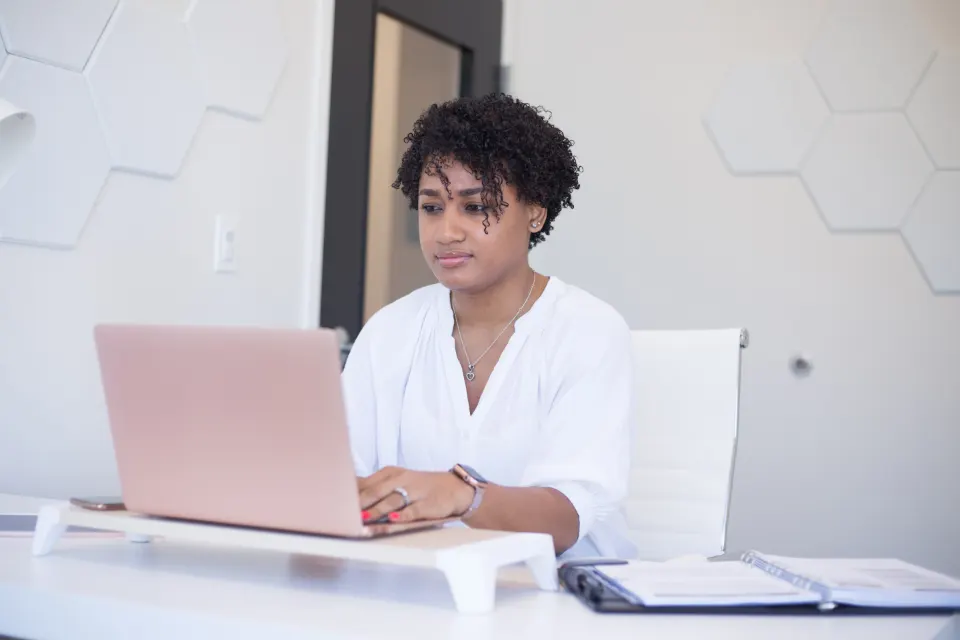 Mulher de camisa branca digitando em um notebook, sentada a uma mesa branca com um fichário ao lado