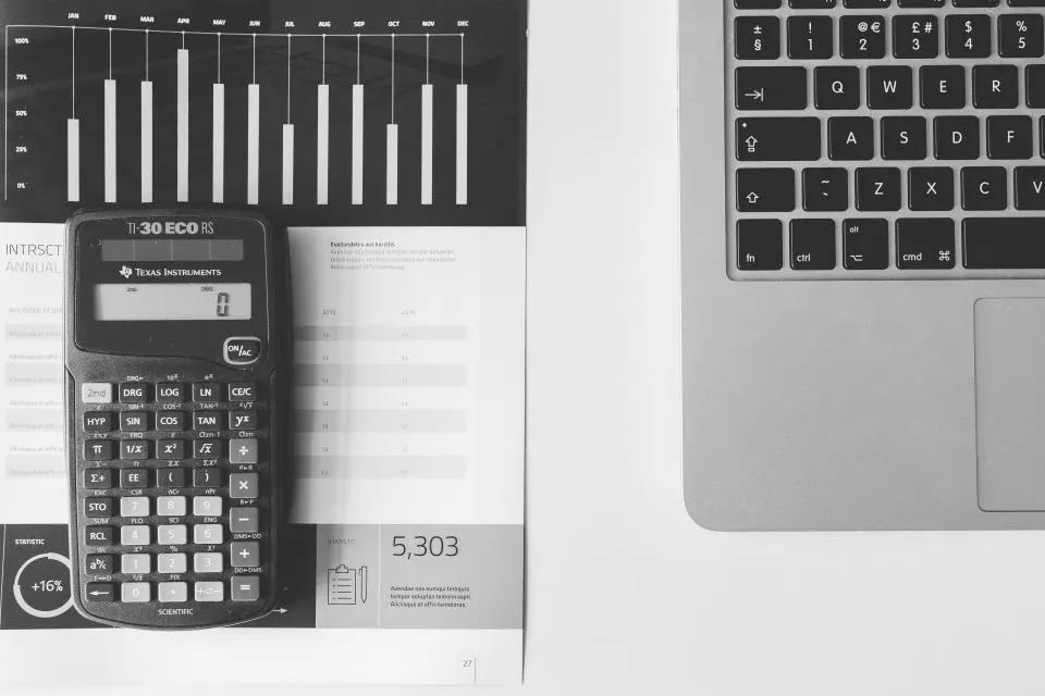 Calculadora sobre um relatório com gráfico de barras, ao lado de um notebook, em uma mesa branca