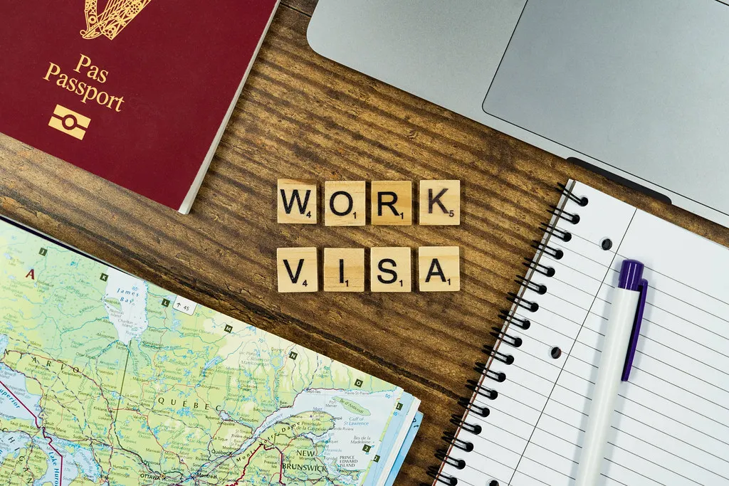 Passaporte, notebook, mapa e caderno sobre uma mesa de madeira, com letras formando as palavras work visa